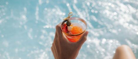 A person holding a cocktail with an olive garnish, with a pool in the background.