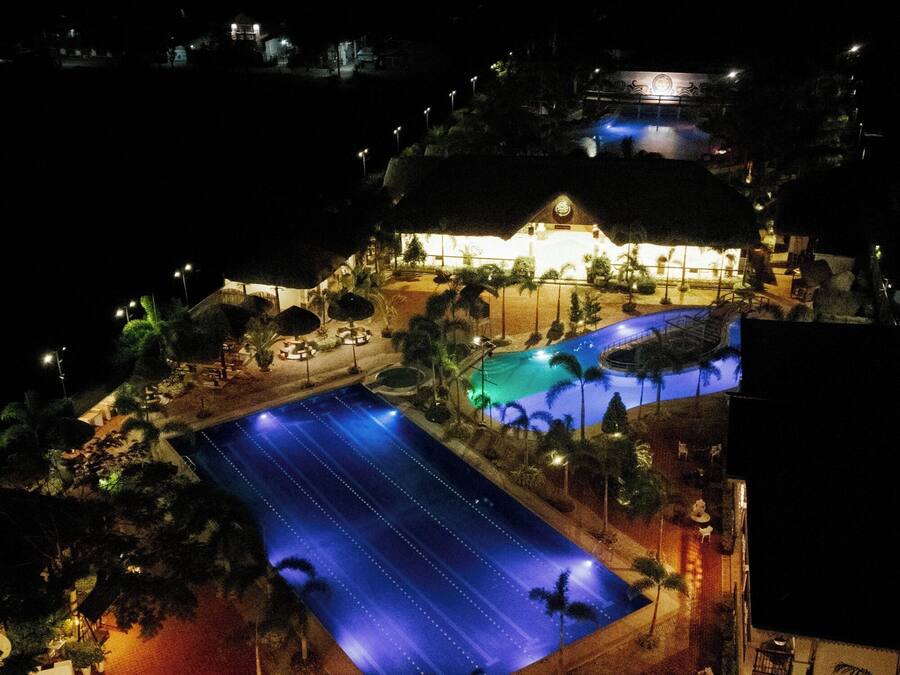 An aerial view of a resort with a large swimming pool, a covered area with seating, and a building with illuminated windows.