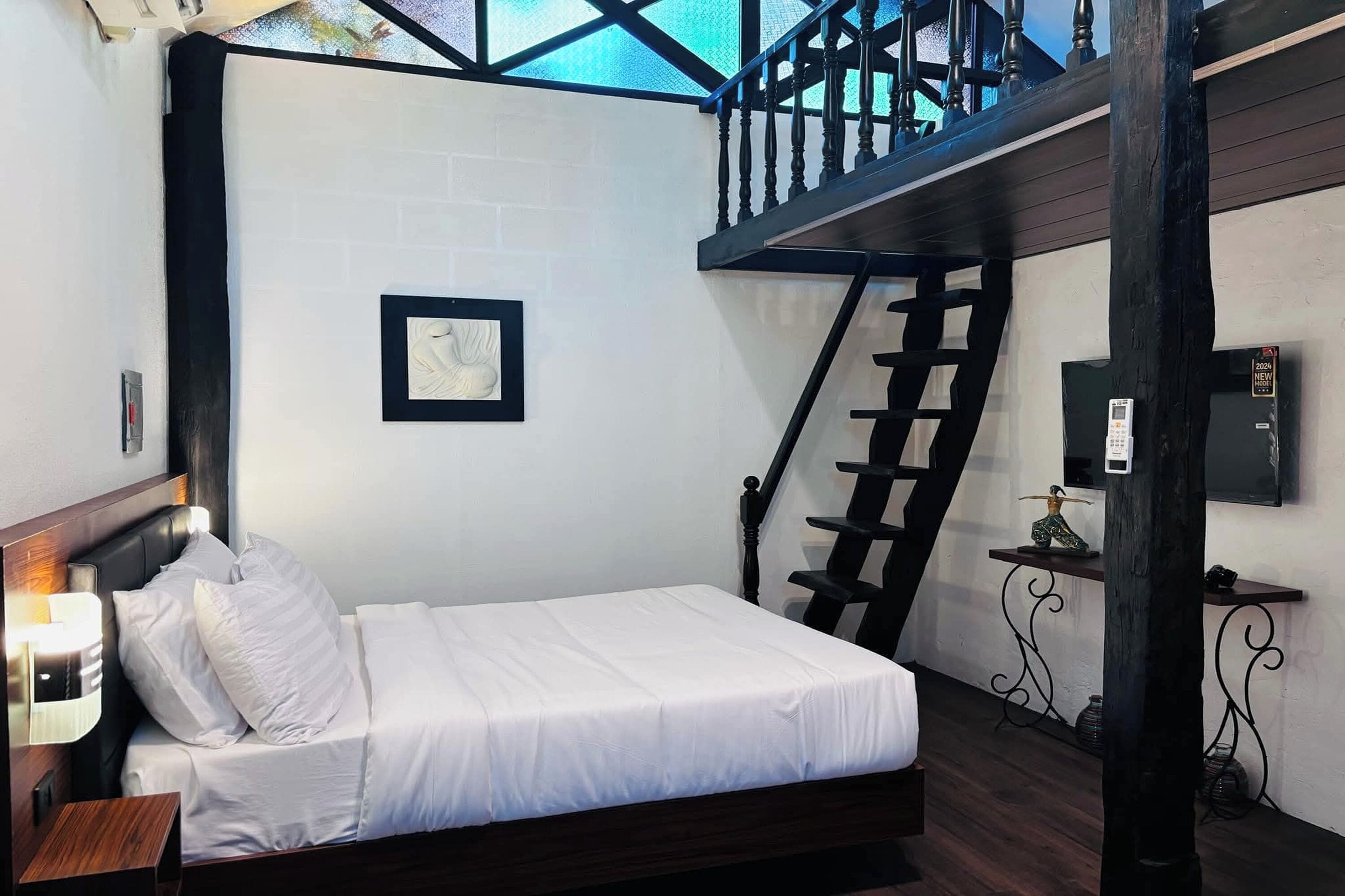 Una cama bien hecha en una habitación con pared blanca, un cuadro enmarcado, cabecera de madera, mesita de noche con lámpara, escalera con barandilla negra y una pequeña mesa con objetos decorativos.