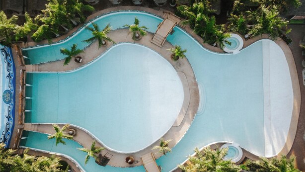 An aerial view of a large, uniquely shaped swimming pool surrounded by palm trees and lounge areas. - Ilocos Urban Wave Resort Hotel (Laoag)