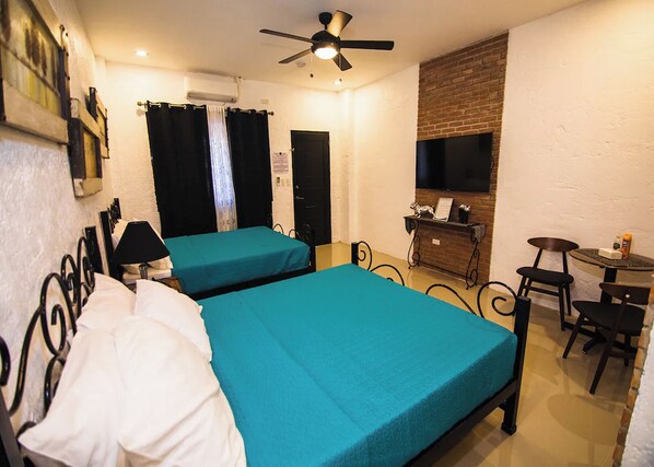 Habitación de hotel con dos camas, ventilador de techo, televisión, una mesita pequeña y pared de ladrillo.