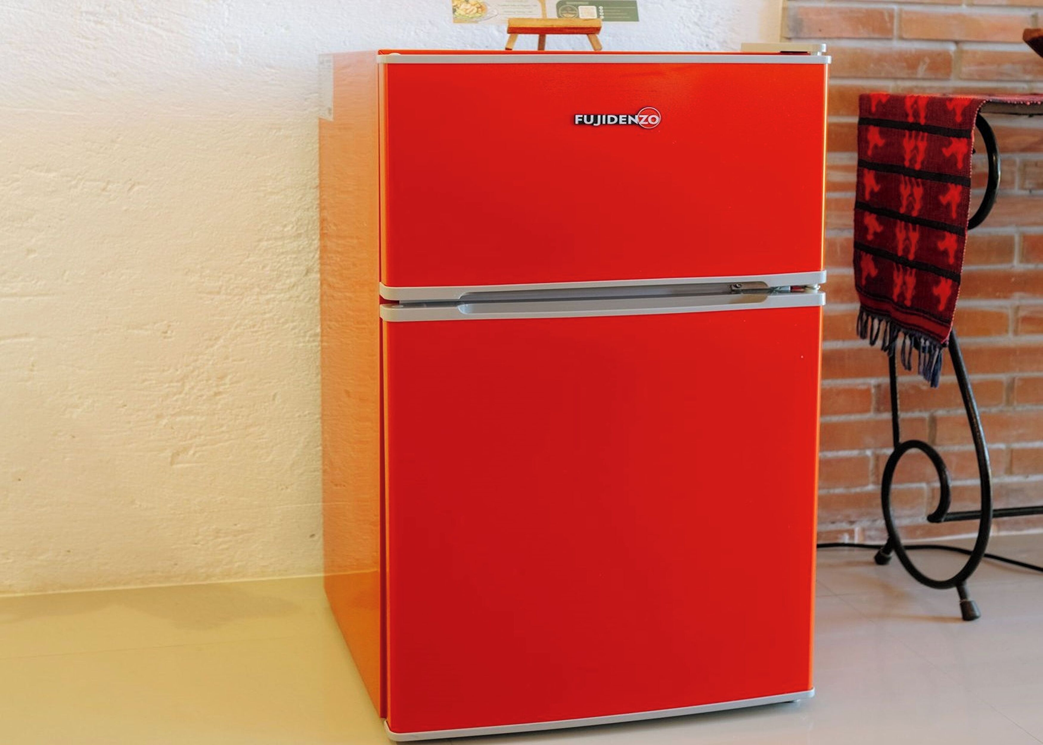 Un refrigerador rojo Fujidenzo en una cocina con pared de ladrillo y un estante metálico con un paño rojo.