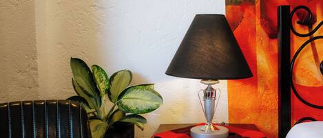 A room with a lamp on a small table, a potted plant, and a bed with a white cover.