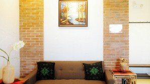 A room with a brick wall, a framed painting, a brown sofa with patterned cushions, a small table with a pineapple decoration, and a vase with flowers. - Ilocos Urban Wave Resort Hotel (Laoag)