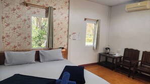 A hotel room with a bed, two windows with curtains, a small table with a chair, and an air conditioning unit on the wall.