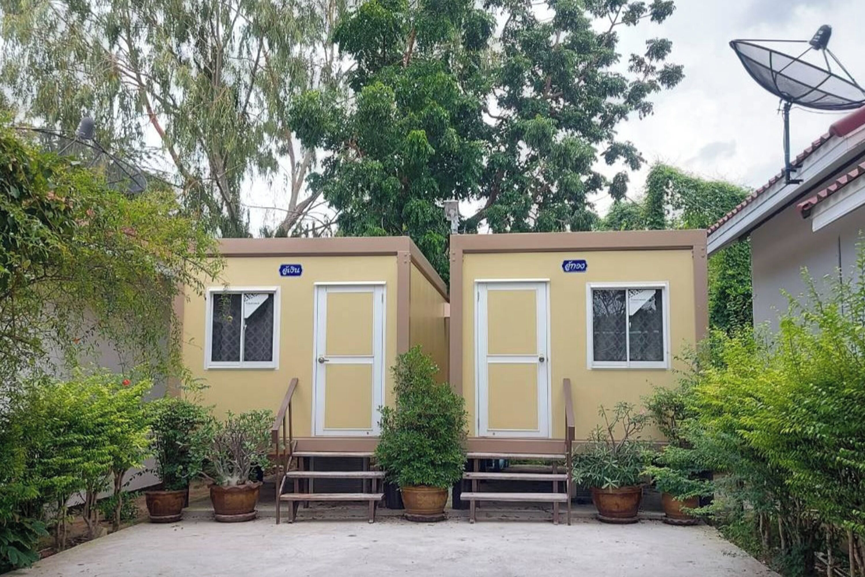 Two small, single-story buildings with white doors and windows, surrounded by potted plants and trees.
