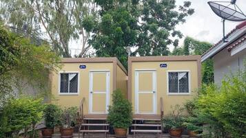 Two small, single-story buildings with white doors and windows, surrounded by potted plants and trees.
