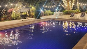 A swimming pool with a tiled edge, surrounded by a garden with various plants and a pergola with string lights.