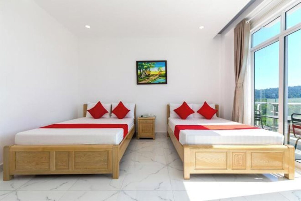 A hotel room with two beds, each with red pillows and white bedding, a small bedside table, a painting on the wall, and a large window offering a view.