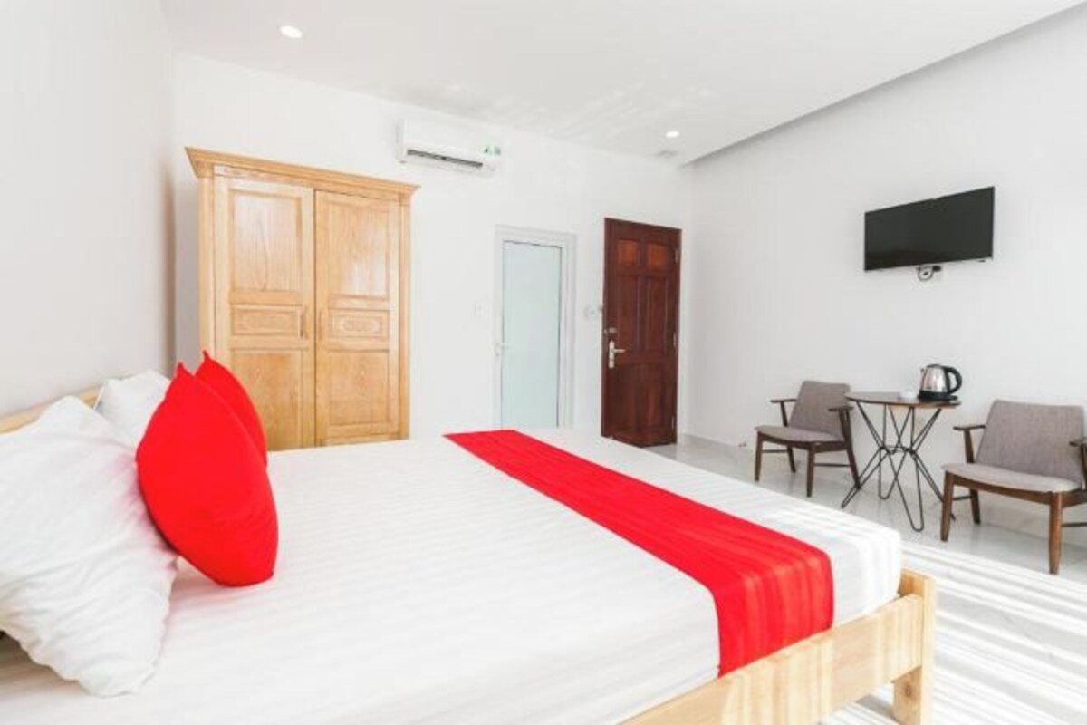 A hotel room with a bed, a wooden wardrobe, a desk with chairs, and a television on the wall.