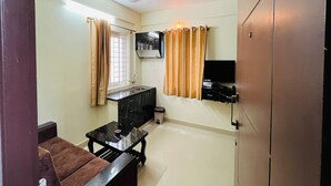 A compact living space with a kitchenette, a ceiling fan, a television mounted on the wall, and a window with curtains. - Marvel Comforts Near Fortis Hospital, Bannerghatta Road (Bengaluru)