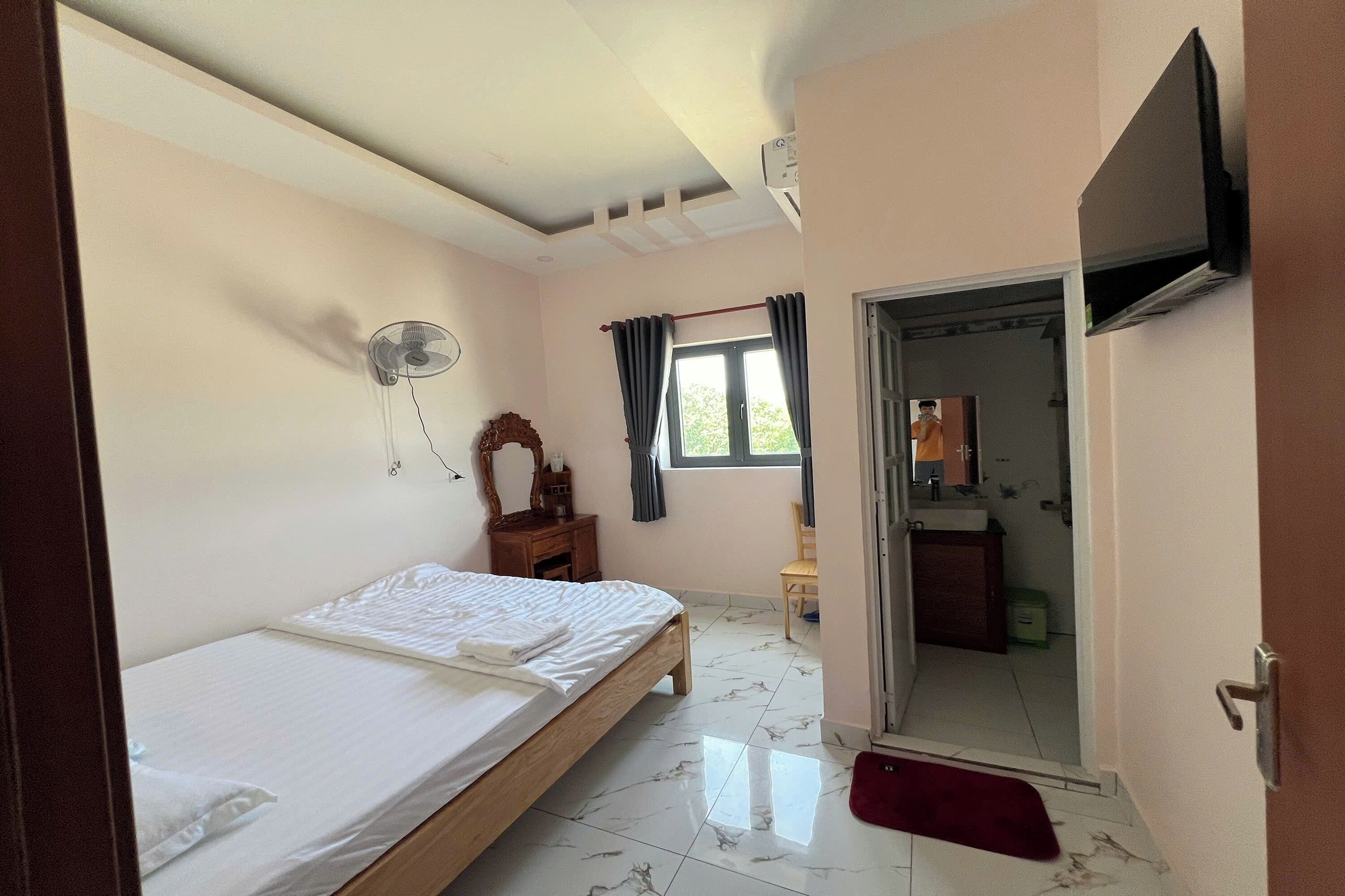 Un dormitorio con cama, ventilador, ventana con cortinas, televisor montado en la pared y puerta que da a un baño.