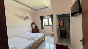 Un dormitorio con cama, ventilador, ventana con cortinas, televisor montado en la pared y puerta que da a un baño.