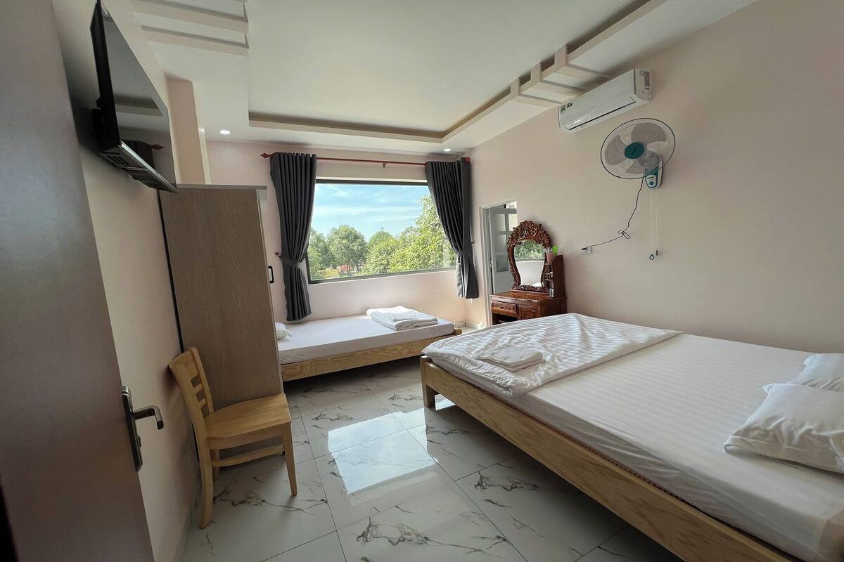 A hotel room with a bed, a chair, a window with curtains, a TV, an air conditioner, and a mirror.
