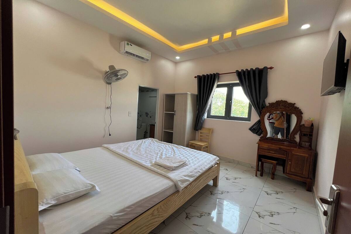 A bedroom with a bed, bedside table, dresser, vanity, mirror, and an air conditioning unit.