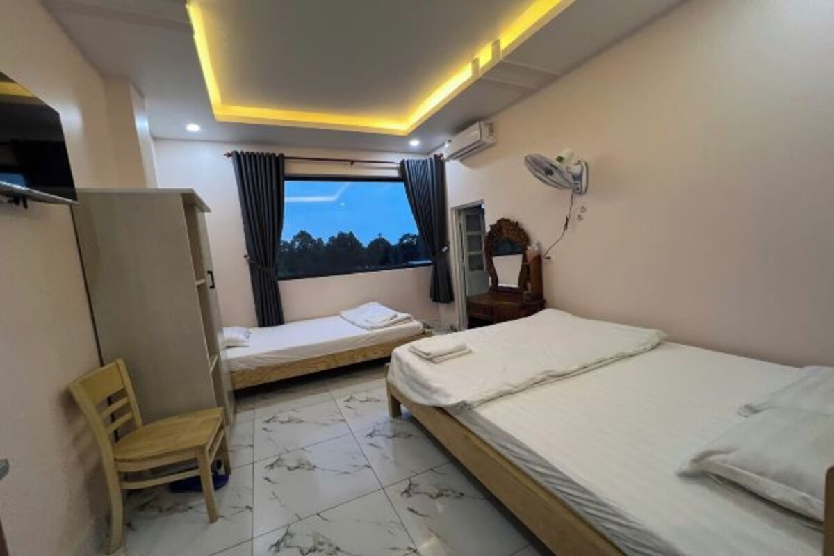 Une chambre avec deux lits simples, une chaise en bois, une armoire, une fenêtre avec des rideaux et un ventilateur de plafond.