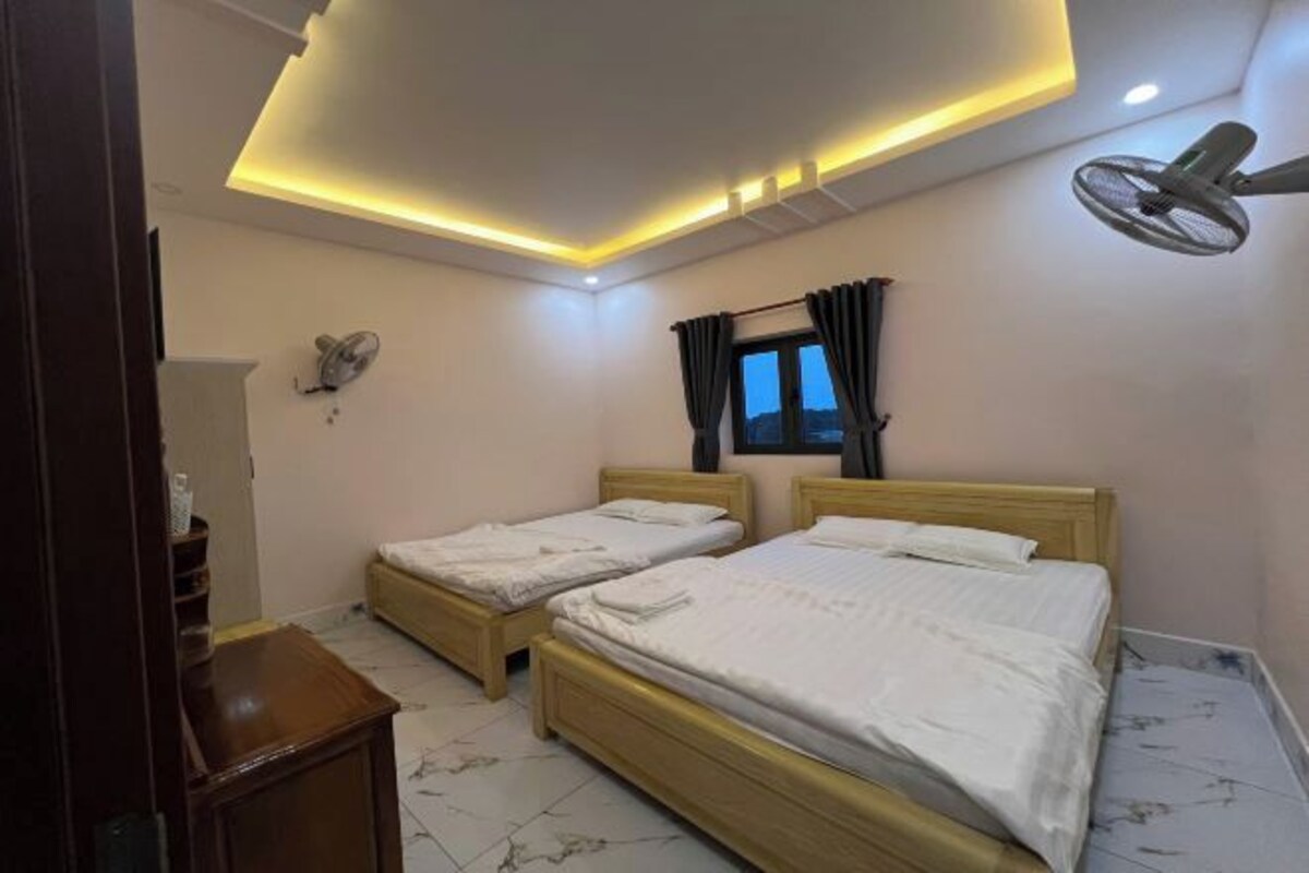 Habitación de hotel con dos camas individuales, un ventilador de techo, una ventana con cortinas y un ventilador fijado a la pared.