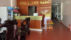 A hotel reception area with a wooden reception desk, a fan, a refrigerator, and a decorative vase with flowers. - Thanh Hang Hotel (Ho Chi Minh City)