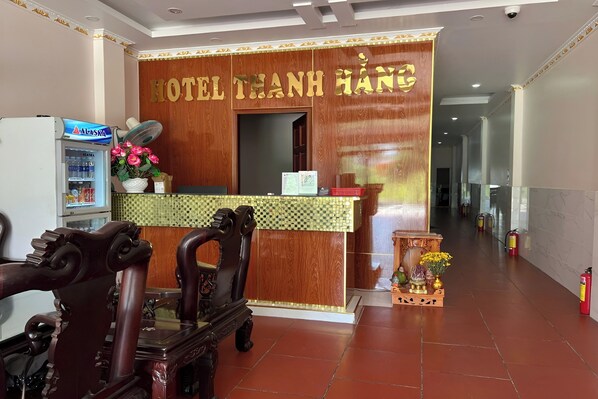 Zona de recepción de un hotel con un mostrador de madera, un ventilador, un refrigerador y un jarrón decorativo con flores.