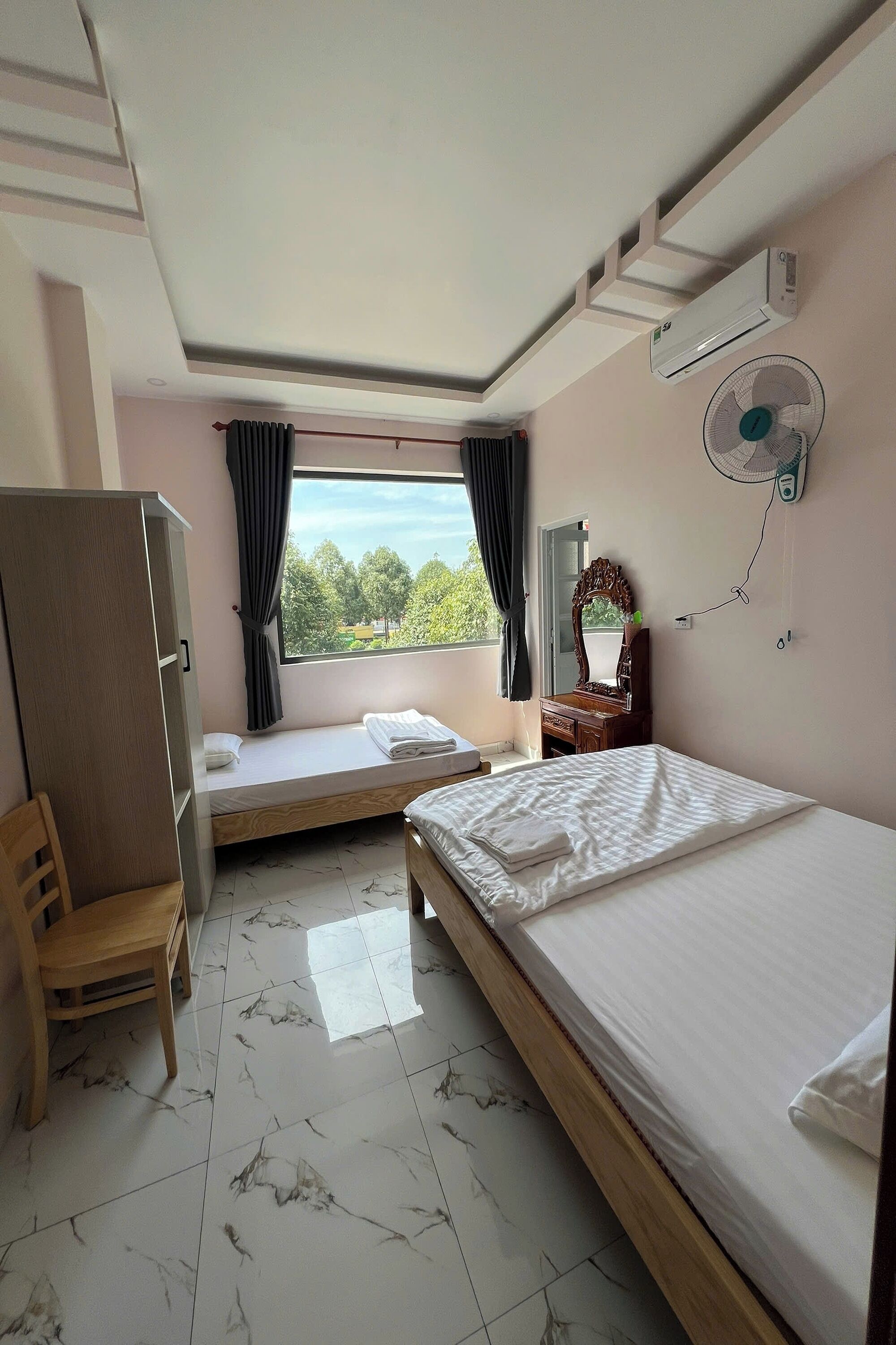 A bedroom with two beds, a wardrobe, a mirror, a window with curtains, and a ceiling fan.