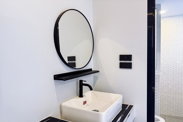 A modern bathroom with a white sink, black faucet, and a round mirror. There is a black shelf above the sink and a black electrical outlet on the wall. - Smith Hotel (Chiang Mai)