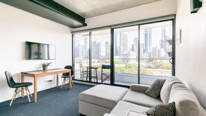 Ein modernes Wohnzimmer mit einer Couch, einem Esstisch und Stühlen. Ein Fernseher ist an der Wand befestigt, und es gibt ein großes Fenster mit Blick auf die Stadt.