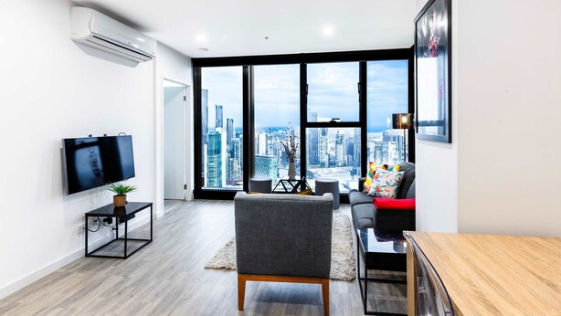 A modern living room with a large window offering a city view, a flat-screen TV, a grey sofa, and a wooden dining table.