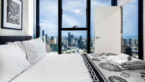 A hotel room with a large window offering a city view, a bed with white linens, and a framed picture on the wall.