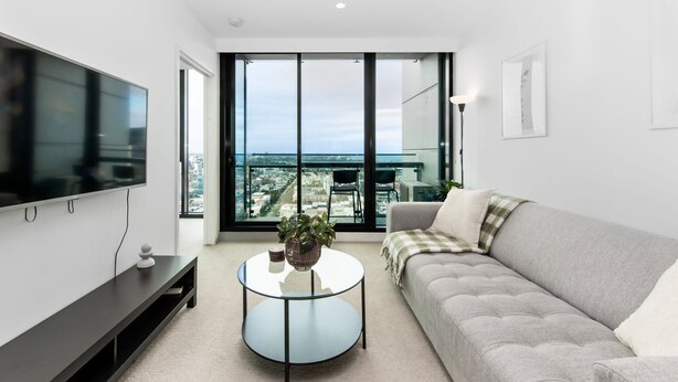 A modern living room with a large flat-screen TV, a round glass coffee table, a grey sofa, and a view of the city through floor-to-ceiling windows.