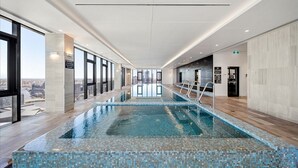 A modern indoor swimming pool with clear glass walls, a tiled floor, and a city view.