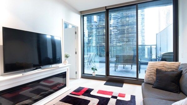 A modern living room with a flat-screen TV, a fireplace, a sofa, and a large sliding glass door leading to an outdoor balcony with a city view.