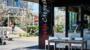 Un escaparate con un letrero negro que dice "Cravin' Crepes" y un cartel de 'cerrado' en la ventana. Hay mesas y sillas en la calle.