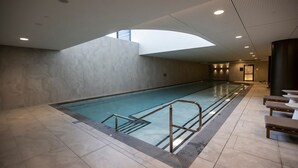 A large indoor swimming pool with a modern design, surrounded by a tiled floor and walls, and a seating area with tables and benches.