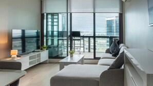 A modern living room with a sofa, a TV, a small table, and a balcony with a view of buildings.