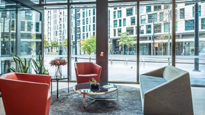 A modern hotel lobby with large windows, red and grey chairs, a glass coffee table, and a view of the cityscape.