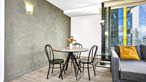En moderne stue med et rundt spisebord, sorte stole, en grå sofa med en gul pude og udsigt til byen gennem store vinduer.