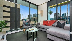 A modern living room with a grey sofa, a patterned armchair, and a small round table. Large windows offer a view of the city skyline and construction cranes.