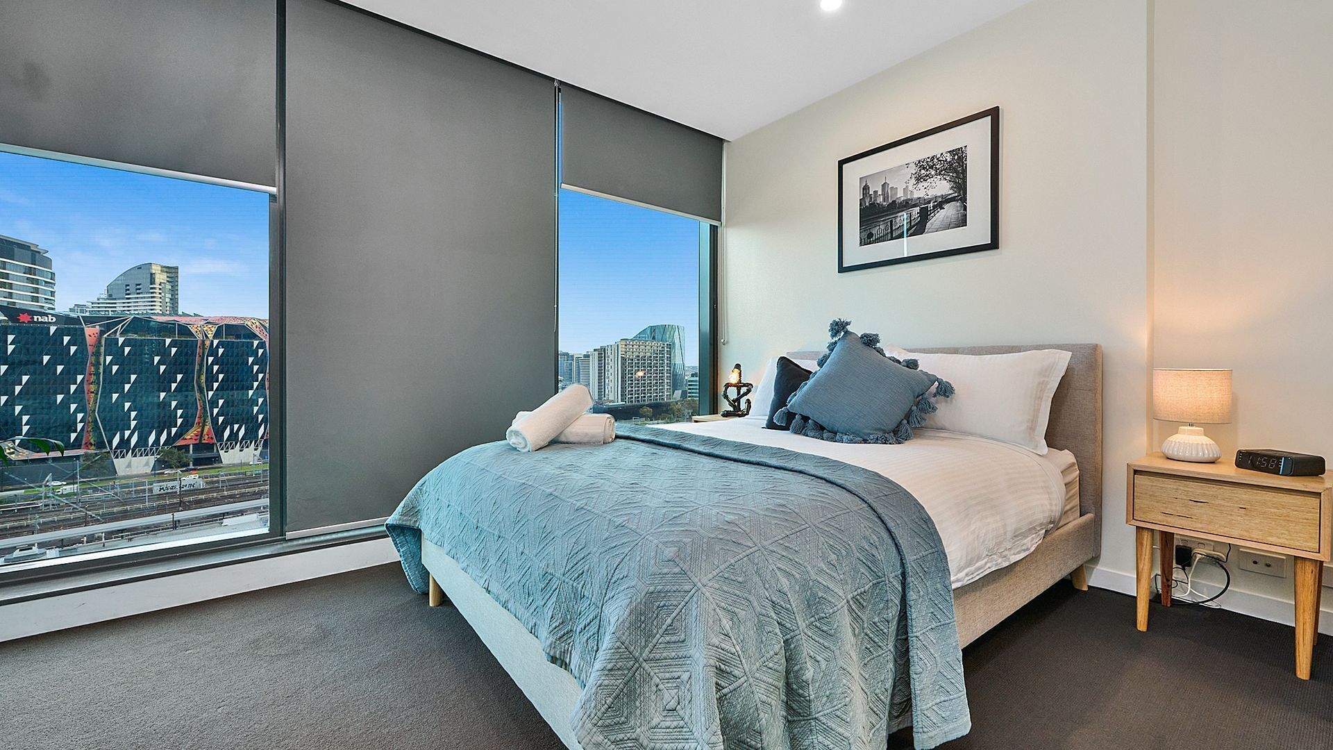 A modern bedroom with a large bed, a nightstand, a framed picture, and a view of a cityscape through large windows.