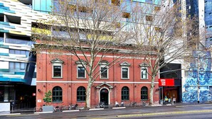 A red brick building with arched windows and a black door, located in an urban area with modern buildings and a mural on the right.