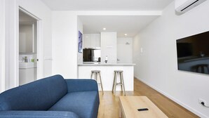A modern living room with a blue sofa, a wooden coffee table, and two stools. A kitchen is visible in the background with white cabinets and a large window.