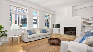 Un salón amplio con un sofá blanco, una mesa de centro y una chimenea. Grandes ventanales ofrecen vistas del paisaje urbano.