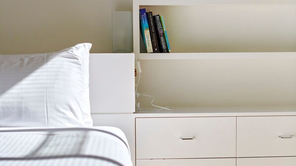Una cama bien hecha con ropa blanca, una mesita de noche blanca con cajón y una estantería con varios libros.