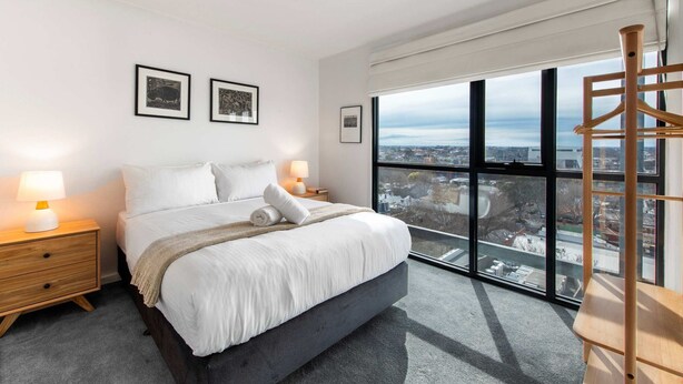 A modern bedroom with a large bed, two bedside tables, a view of the city through large windows, and framed artwork on the wall. - Sweet Escape - Chocolate Factory (Fitzroy)