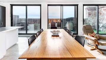 A modern dining area with a wooden table, black chairs, and a view through large glass doors to an outdoor space with a brick wall and greenery.
