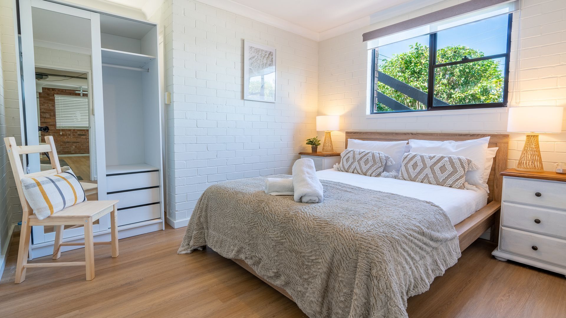 Une chambre à coucher comprenant un lit, une chaise, une table de chevet, une lampe et une fenêtre donnant sur des arbres.