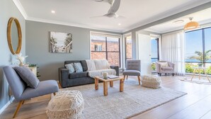 A spacious living room with a grey sofa, two armchairs, a coffee table, and a large window offering a view of a brick building and greenery. - Spectacular Ocean View Home in Bombo (Bombo)