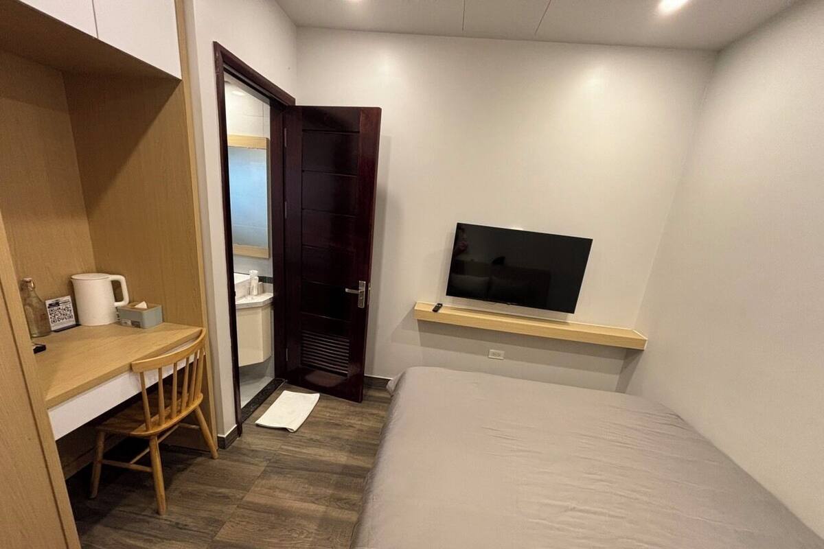 Een kleine, verzorgde slaapkamer met een bed, een bureau met stoel, een televisie op een plank en een kledingkast.