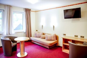 A hotel room with a sofa, two armchairs, a small table, a TV mounted on the wall, and a window with curtains.