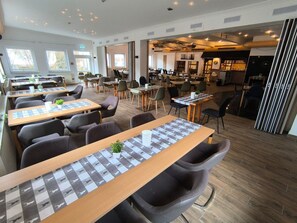 Een modern restaurant met verschillende eetgedeeltes, waaronder een lange gedeelde tafel en kleinere tafels met stoelen.