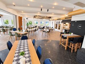 A modern dining area with multiple tables and chairs, large windows, and a central decorative element. - Comfortable and versatile room -perfect for relaxation, work and exchange (Winterberg)